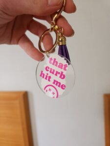 a keychain with a round acrylic tag featuring the humorous phrase that curb hit me in pink text, accompanied by a purple tassel.