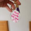 a keychain with a round acrylic tag featuring the humorous phrase that curb hit me in pink text, accompanied by a purple tassel.